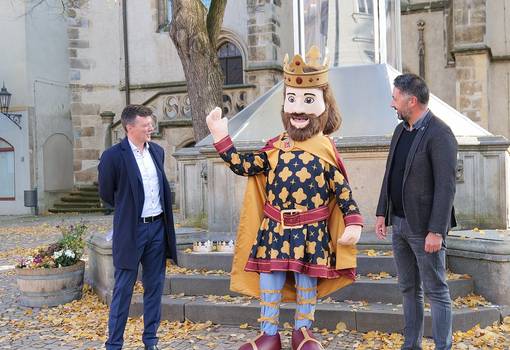 Zwei Männer und in ihrer Mitte ein Königsmaskottchen vor Brunnen