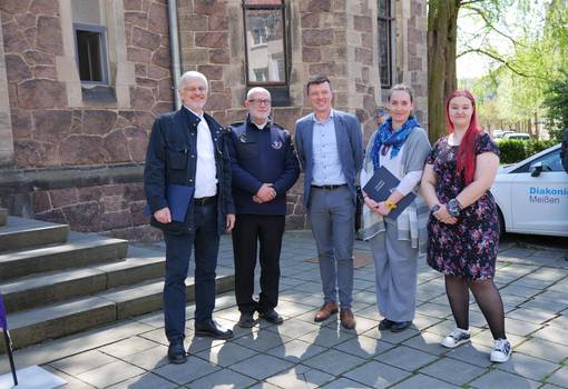 5 Personen vor Treppenstufen und einer Kirche stehen auf grauem Untergrund