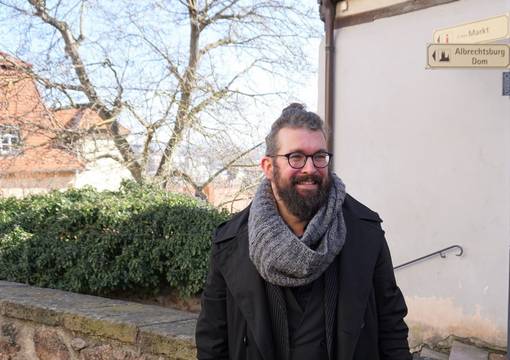Mann Mitte 30 mit Bart, Brille und schwarzem Mantel schaut lächelnd aus dem Bild