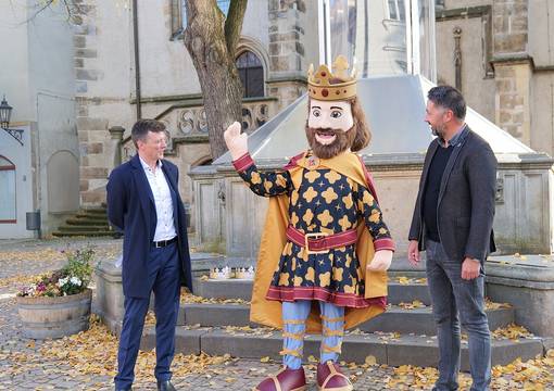 Zwei Männer und in ihrer Mitte ein Königsmaskottchen vor Brunnen