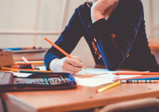 Kind mit dunklem Pullover sitzt an braunem Schreibtisch mit Federmappe, Stiften und Papier darauf