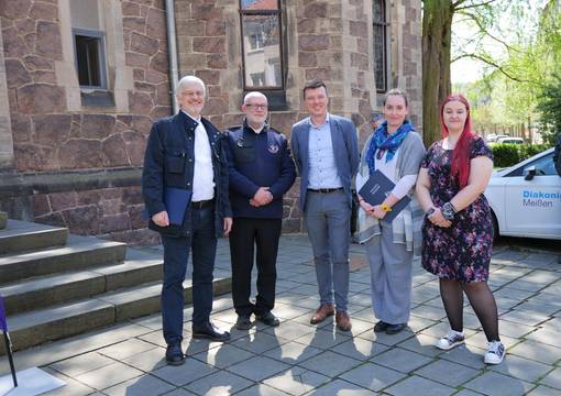 5 Personen vor Treppenstufen und einer Kirche stehen auf grauem Untergrund