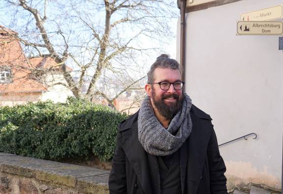 Mann Mitte 30 mit Bart, Brille und schwarzem Mantel schaut lächelnd aus dem Bild