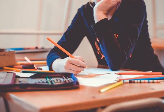 Kind mit dunklem Pullover sitzt an braunem Schreibtisch mit Federmappe, Stiften und Papier darauf